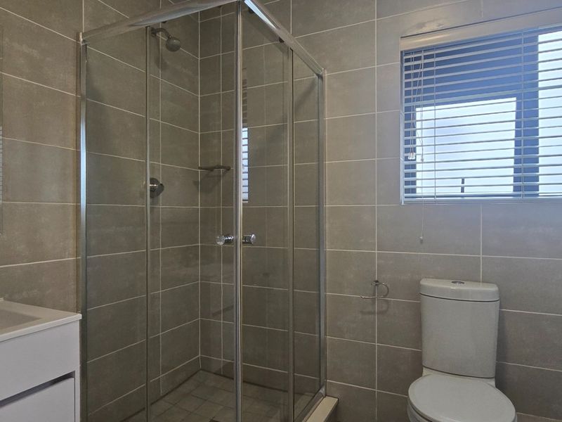 A modern, well-maintained bathroom featuring a glass shower enclosure, vanity basin, toilet, and neutral wall tiling with natural light from a window.