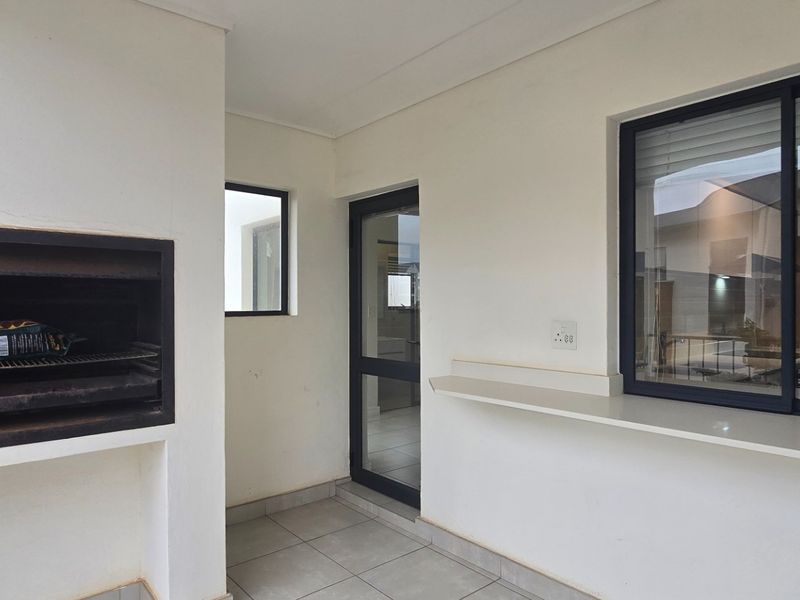 A clean, covered patio area featuring a built-in braai, tiled flooring, and direct access to the interior through a glass door.