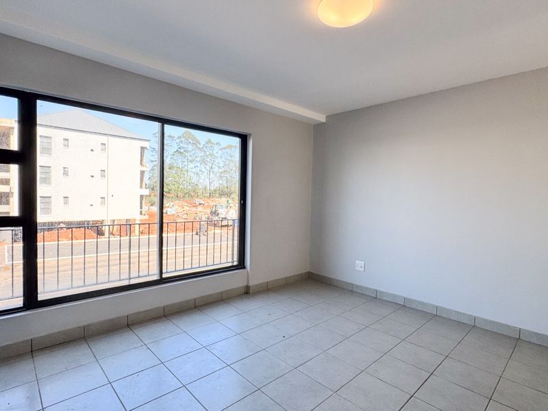 A bright, empty bedroom featuring neutral tiled flooring, clean walls, and a large sliding window providing natural light and an exterior view.