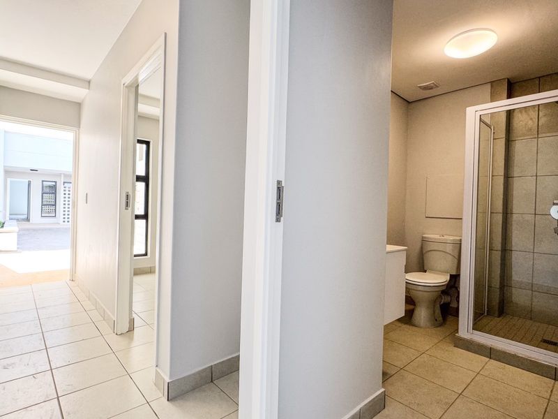 A bright hallway leading to a bathroom featuring a shower enclosure, toilet, and vanity cabinet, with neutral tiled flooring throughout.