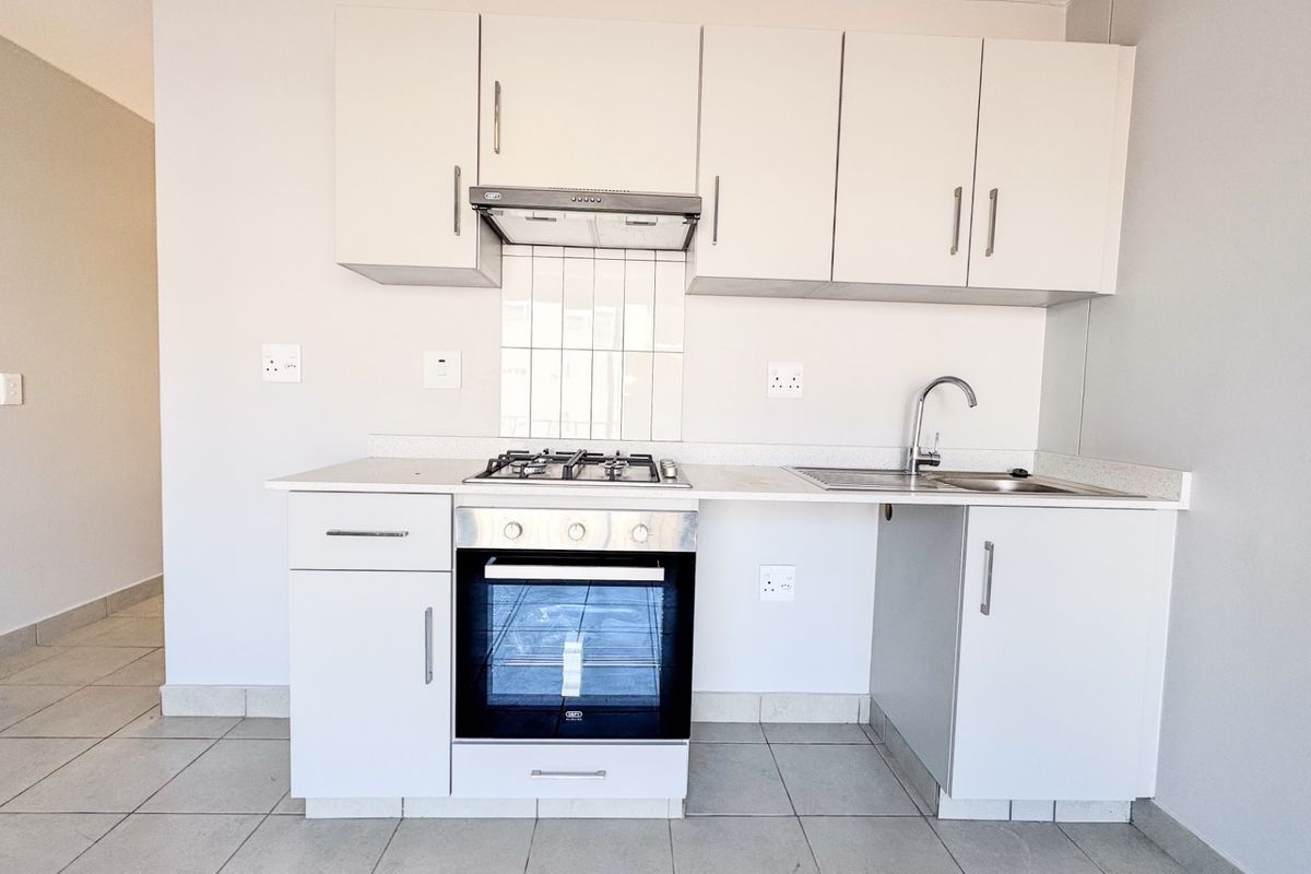 A clean, modern kitchen featuring white cabinetry, a gas hob, built-in oven, extractor fan, and stainless steel sink with tiled splashback.
