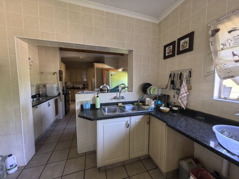 A functional kitchen featuring granite countertops, double bowl sink, tiled splashbacks, and tiled flooring, with an open-plan layout connecting to an adjacent prep area.