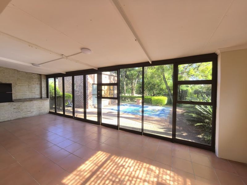 Spacious covered patio featuring tiled flooring, a built-in braai with stone cladding, and large sliding glass doors overlooking the garden and pool area.
