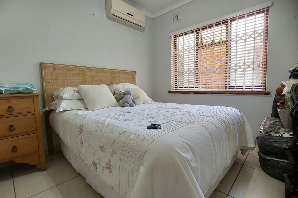 A functional bedroom featuring tiled flooring, a wall-mounted air conditioning unit, and a window fitted with security bars.