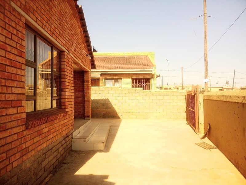 A rear exterior view of a face brick house featuring a concrete patio area, perimeter wall, and metal security gate access.