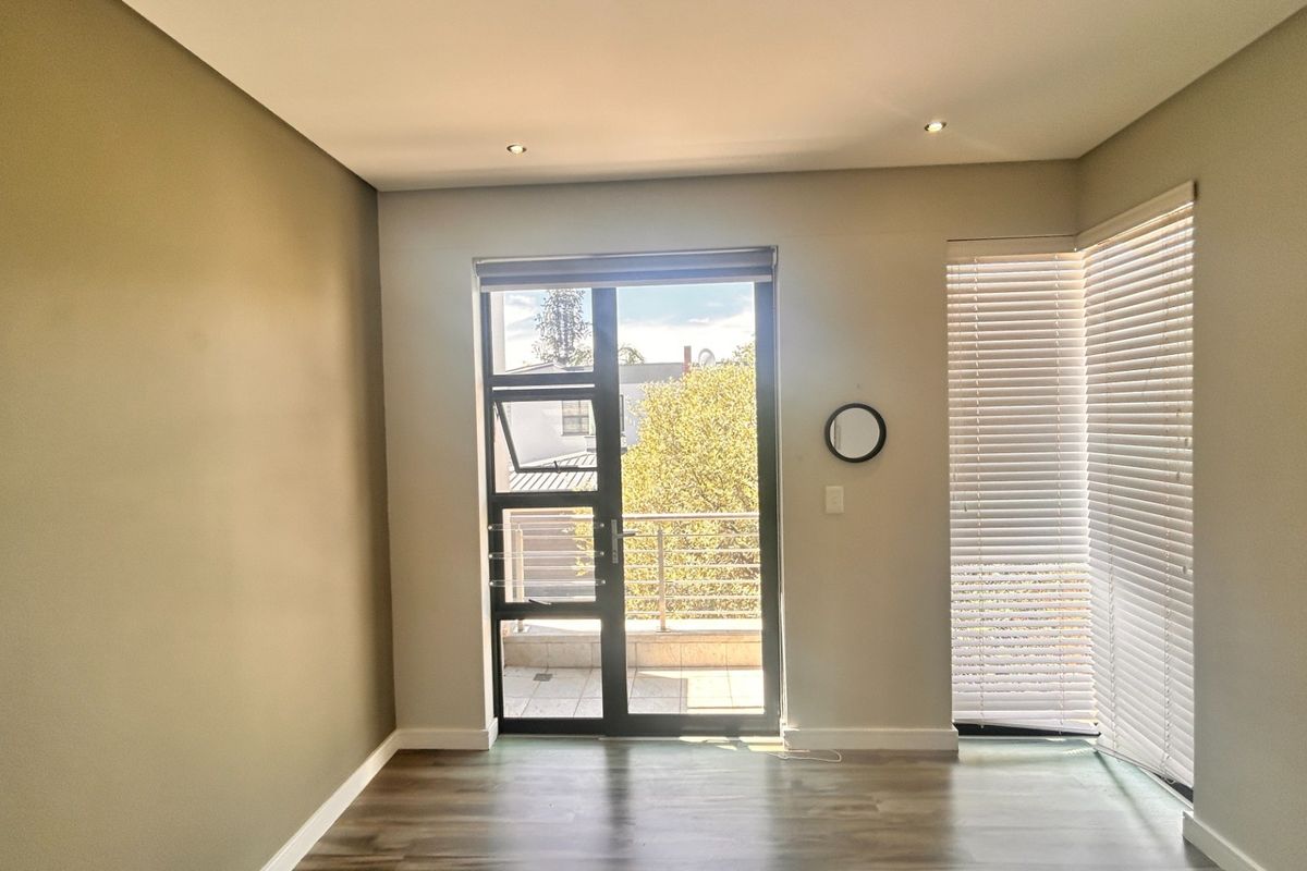 A bright, empty bedroom featuring modern laminate flooring, recessed downlights, and a sliding door leading to a private balcony with venetian blinds.