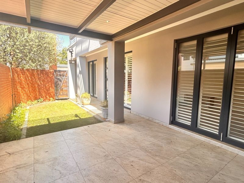 A spacious covered patio featuring tiled flooring, recessed lighting, and large aluminium-framed glass doors leading into the home, overlooking a small private garden.