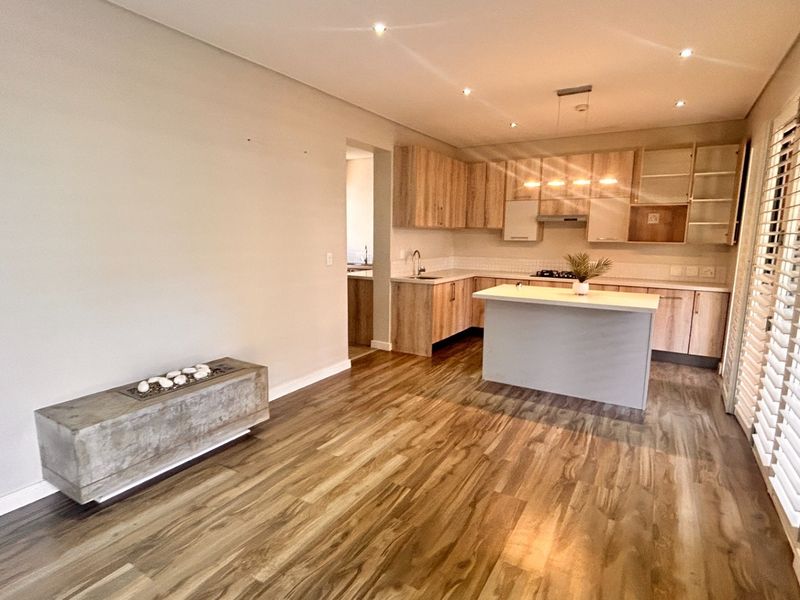 Spacious open-plan living and kitchen area featuring wood-look flooring, modern cabinetry, a central island, built-in fireplace, and plantation shutters.
