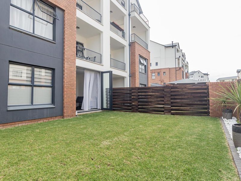 A private rear garden area featuring a well-maintained lawn, decorative pebble border, and timber fencing, adjacent to the apartment building's ground floor patio.