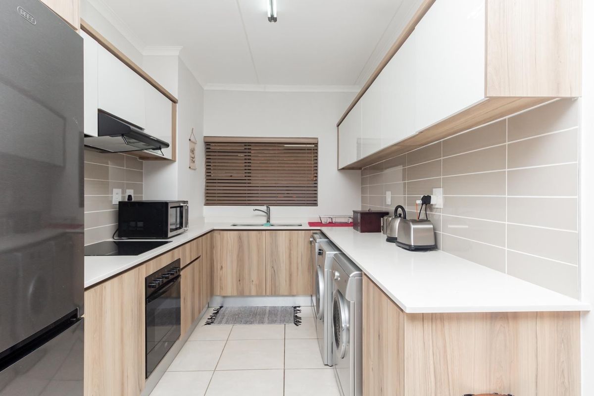 Modern galley-style kitchen featuring light wood-grain cabinetry, white stone countertops, tiled backsplash, and integrated appliances including an oven and hob.