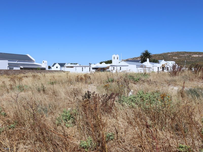A view of a vacant residential plot with dry vegetation, featuring a boundary wall and surrounding white-painted coastal style properties in the background.
