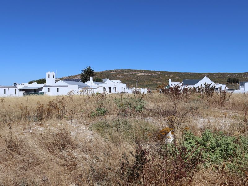 A view of a vacant residential plot featuring dry natural vegetation with established white-walled residential buildings visible in the background.