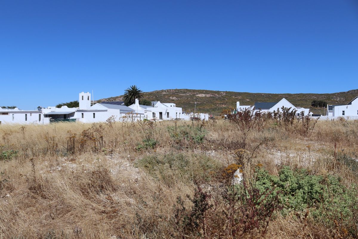 A view of a vacant residential plot featuring dry natural vegetation with established white-walled residential buildings visible in the background.