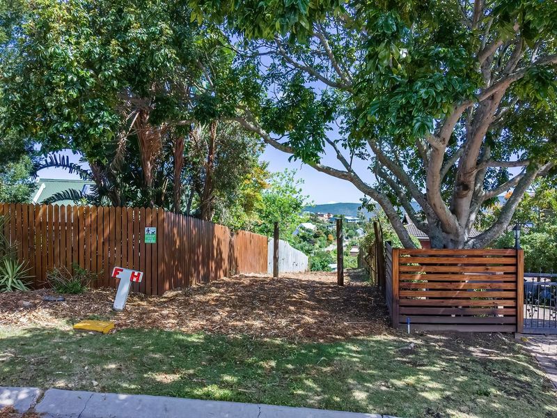A view of the vacant residential plot featuring established trees, wooden perimeter fencing, and a paved driveway entrance.