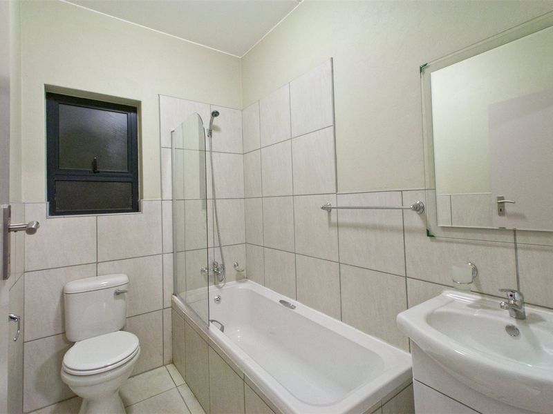 A clean, modern bathroom featuring a bathtub with a glass shower screen, a vanity basin, and neutral wall tiling.