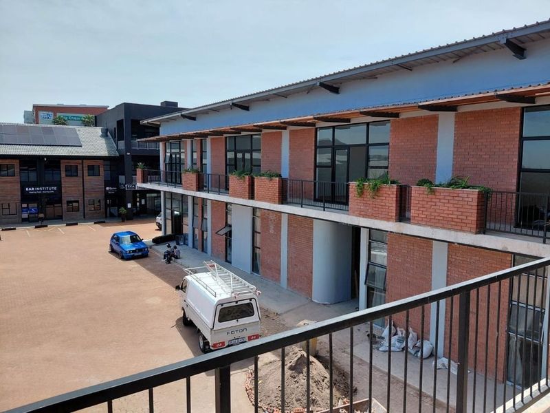 Exterior view of a modern commercial building featuring exposed brickwork, a metal roof, and a balcony with integrated brick planters overlooking a paved courtyard.