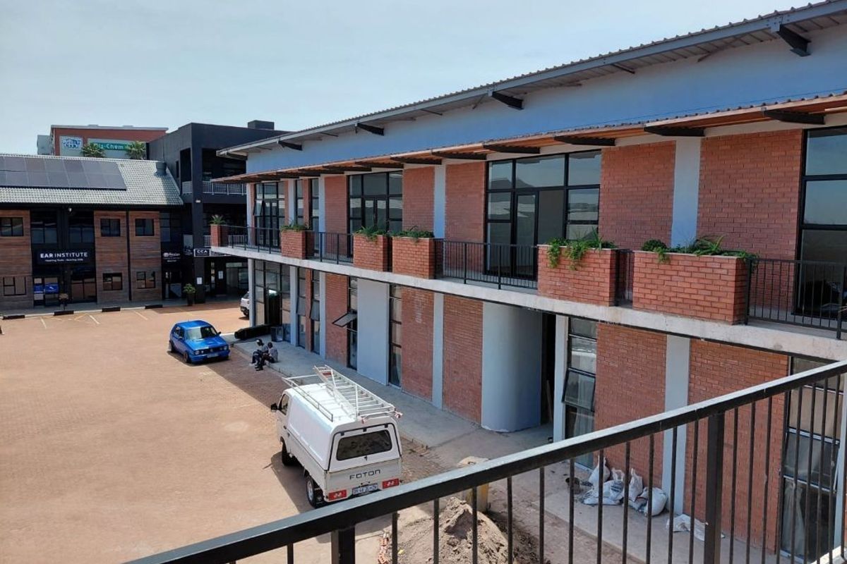 Exterior view of a modern commercial building featuring exposed brickwork, a metal roof, and a balcony with integrated brick planters overlooking a paved courtyard.