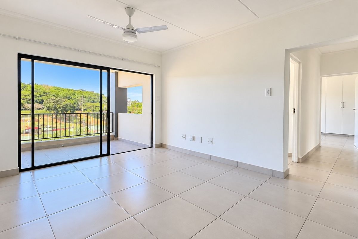 A bright, open-plan living area featuring neutral tiled floors, a ceiling fan, and large sliding doors leading to a private balcony with scenic views.