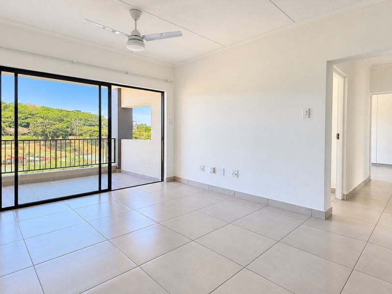 A bright, open-plan living area featuring neutral tiled floors, a ceiling fan, and large sliding doors leading to a private balcony with scenic views.