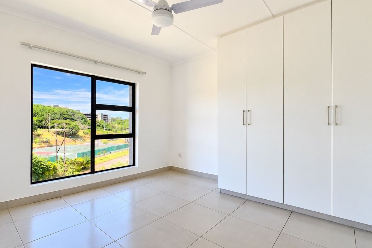 A bright, empty bedroom featuring neutral tiled flooring, built-in white cupboards, a ceiling fan, and a large window with a pleasant outdoor view.