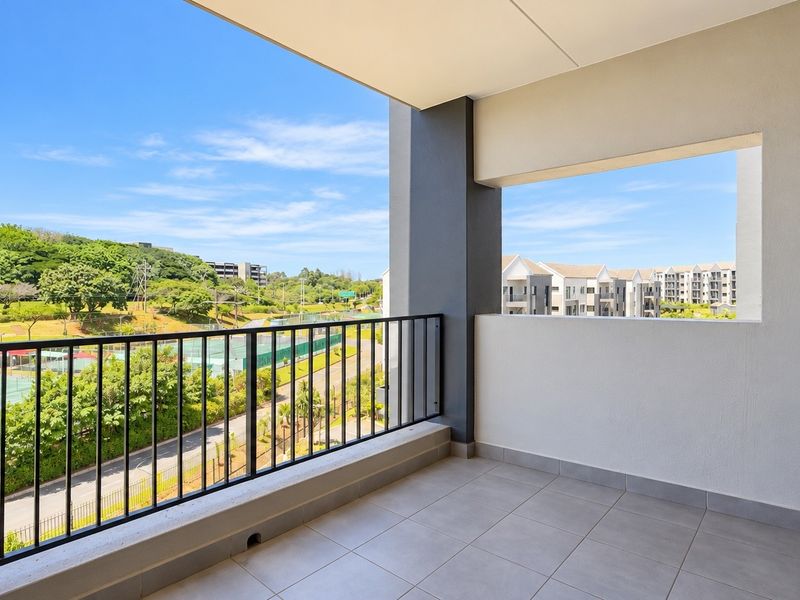 A spacious, modern covered balcony featuring tiled flooring and a metal balustrade, offering elevated views over the surrounding estate and tennis courts.