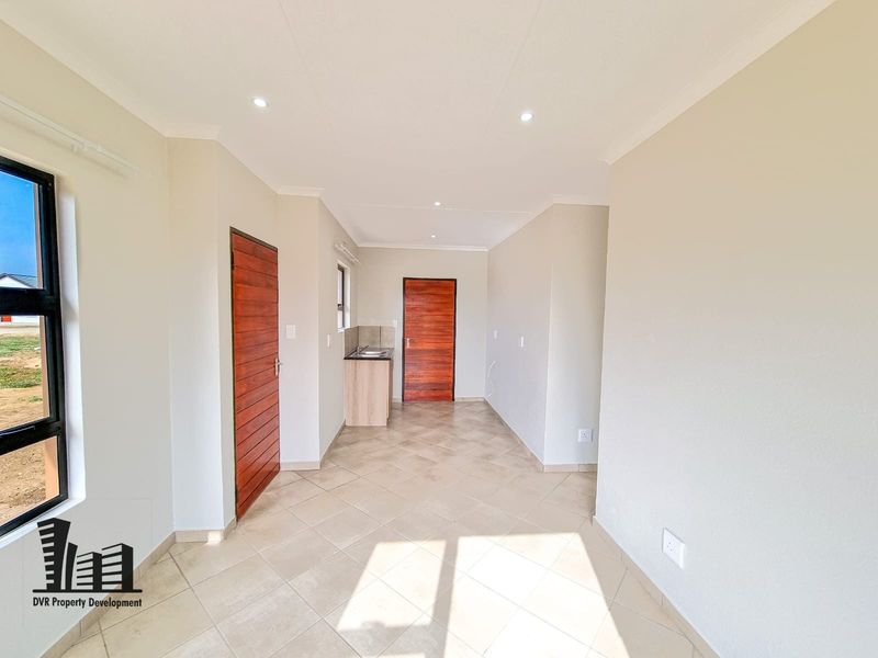 A bright, open-plan living area featuring neutral tiled flooring, recessed lighting, and a compact kitchen corner with a sink unit.