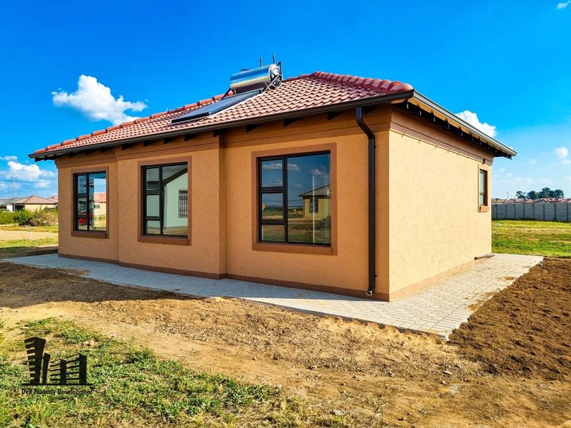 A modern single-story house exterior featuring a tiled roof, solar water heating system, and paved perimeter walkway set in a developing residential area.