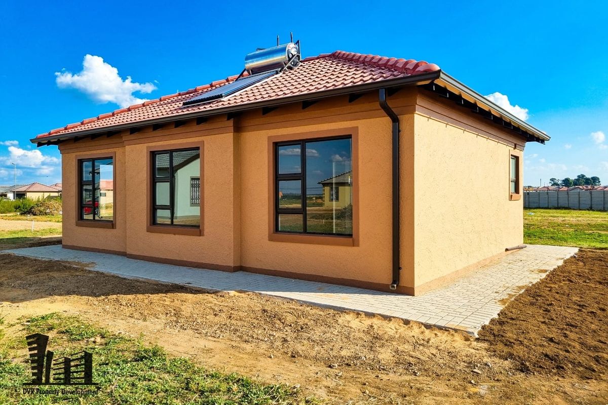 A modern single-story house exterior featuring a tiled roof, solar water heating system, and paved perimeter walkway set in a developing residential area.