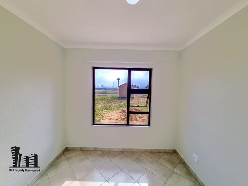 A clean, empty bedroom featuring neutral walls, tiled flooring, and a large aluminium-framed window providing natural light and exterior views.