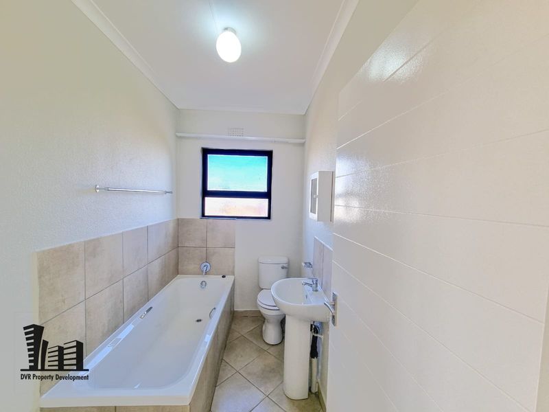 A clean, modern bathroom featuring a white bathtub, pedestal basin, and toilet with neutral tiled finishes and natural light.