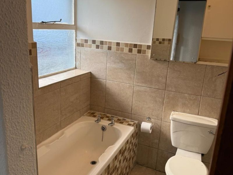 A compact bathroom featuring a bathtub, toilet, and neutral tiled finishes. The space includes a frosted window and a wall-mounted mirror.