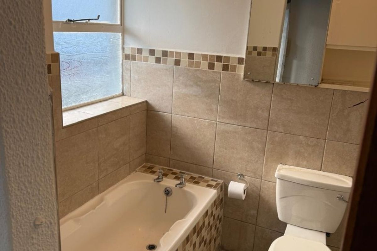A compact bathroom featuring a bathtub, toilet, and neutral tiled finishes. The space includes a frosted window and a wall-mounted mirror.