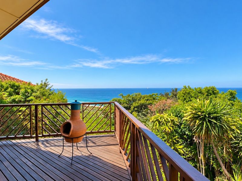 A spacious timber balcony featuring a wooden balustrade and a covered roof section, offering expansive, elevated views of the ocean and surrounding greenery.