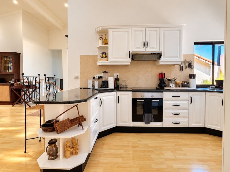 A well-maintained kitchen featuring white cabinetry, granite countertops, built-in oven and hob, and a tiled backsplash, seamlessly integrated into an open-plan living area.