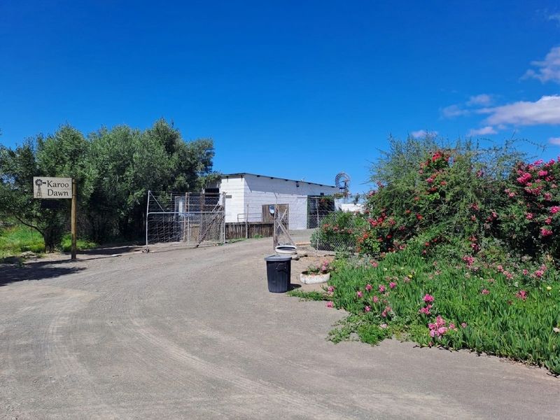 Front approach to the property featuring a gravel driveway, wire fencing, and a traditional windmill structure set against a rural landscape.