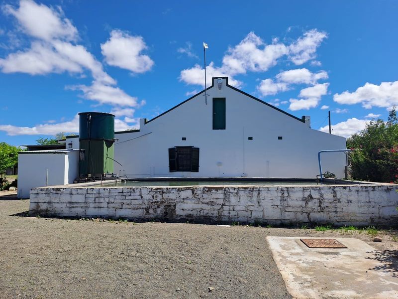 Exterior view of a rustic property featuring a raised stone pool and a large water storage tank against a white gable-end wall.