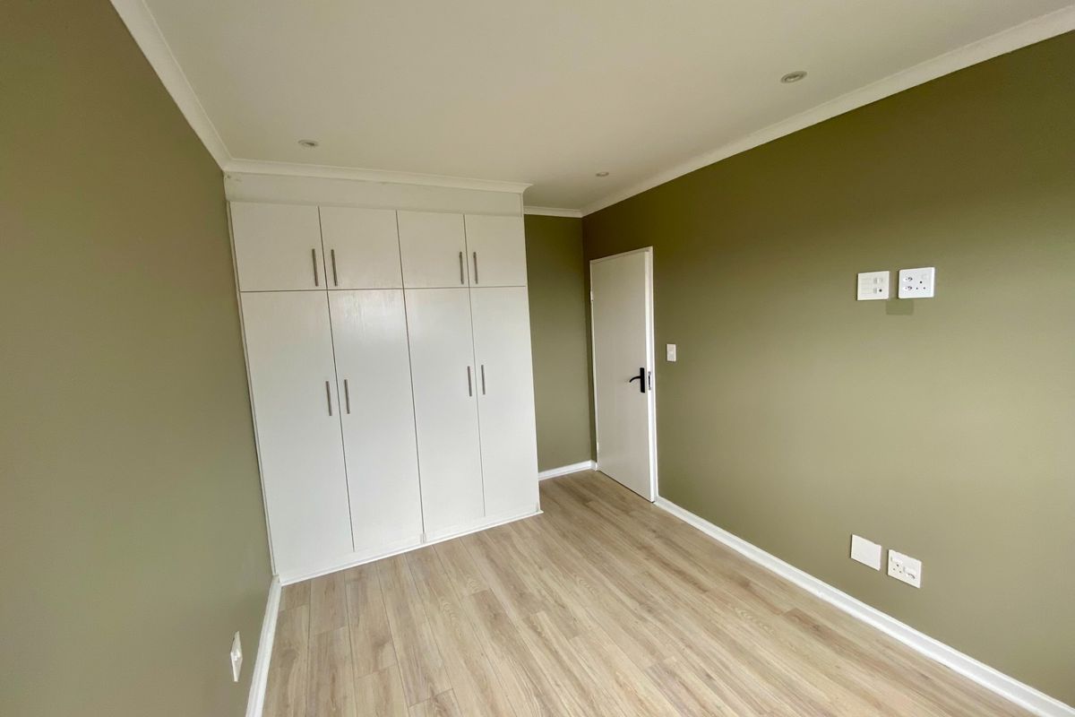 A clean, empty bedroom featuring modern laminate flooring, built-in white cupboards, and neutral olive-toned walls with recessed lighting.
