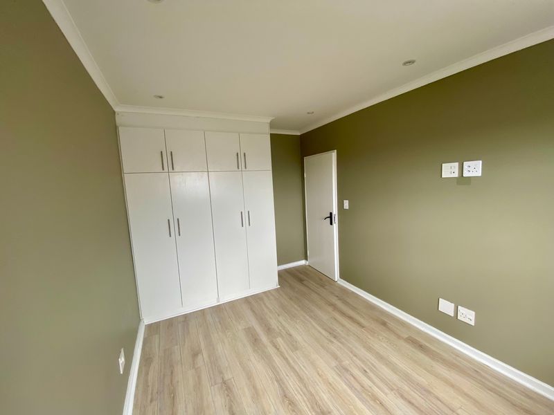 A clean, empty bedroom featuring modern laminate flooring, built-in white cupboards, and neutral olive-toned walls with recessed lighting.