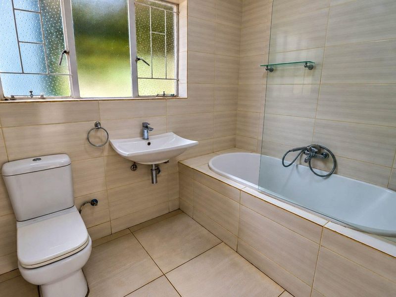A functional bathroom featuring a built-in bathtub with a glass screen, wall-mounted basin, and neutral ceramic tiling throughout.