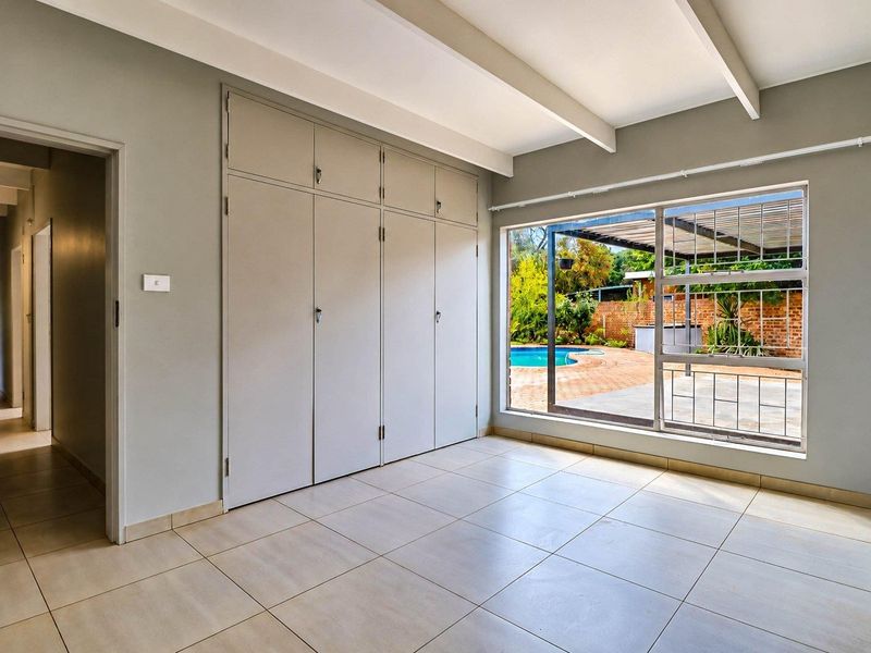 A bright, empty bedroom featuring built-in cupboards, tiled flooring, and exposed ceiling beams, with a view overlooking the outdoor pool area.