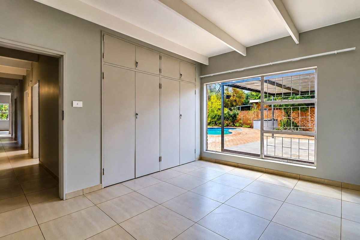 A bright, empty bedroom featuring built-in cupboards, tiled flooring, and exposed ceiling beams, with a view overlooking the outdoor pool area.