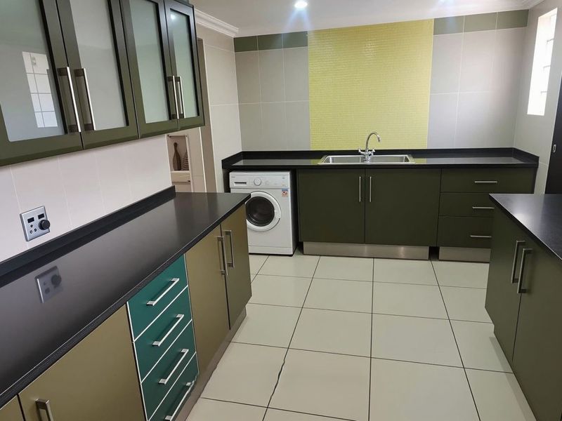 A well-maintained scullery featuring ample built-in cabinetry, dark stone countertops, a stainless steel sink, and integrated laundry facilities with modern tiled finishes.
