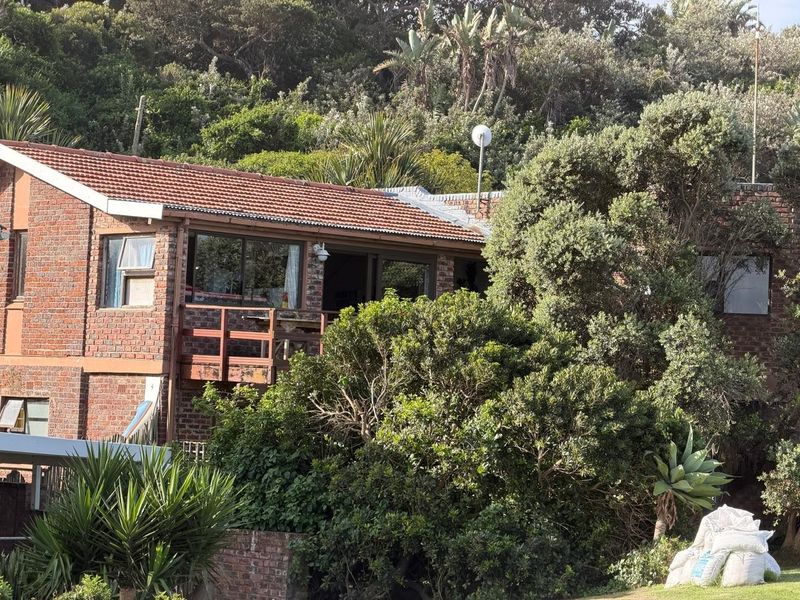 Rear view of a face brick house featuring a terracotta tile roof and a wooden balcony nestled among mature trees and garden foliage.