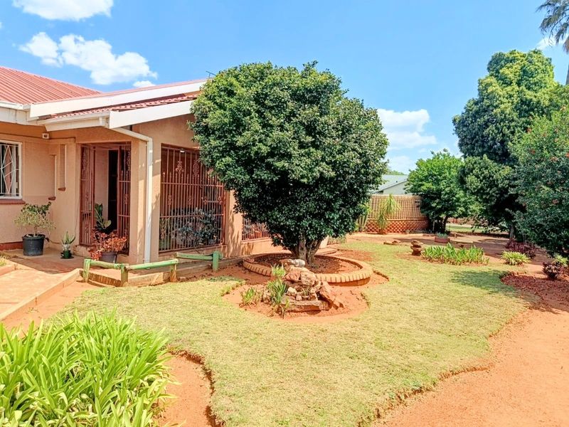 A rear view of the house featuring a covered patio area, metal security gates, and a landscaped garden with established trees and brick-edged beds.