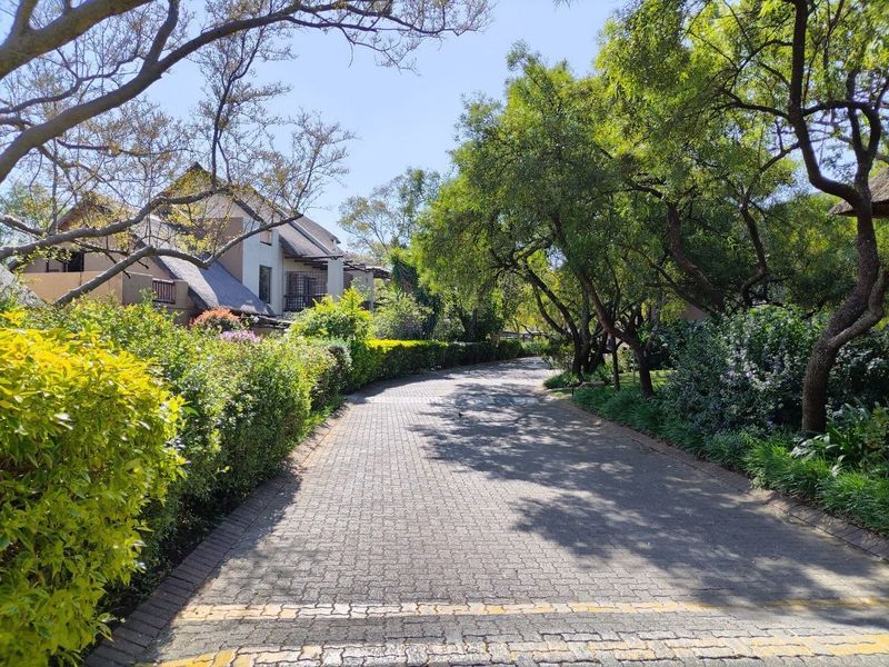 A scenic view of a paved driveway leading towards a townhouse complex with mature trees and well-maintained garden landscaping.