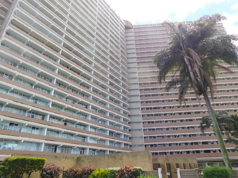 A low-angle exterior view of a large high-rise apartment building featuring multiple levels of balconies and a brick boundary wall.