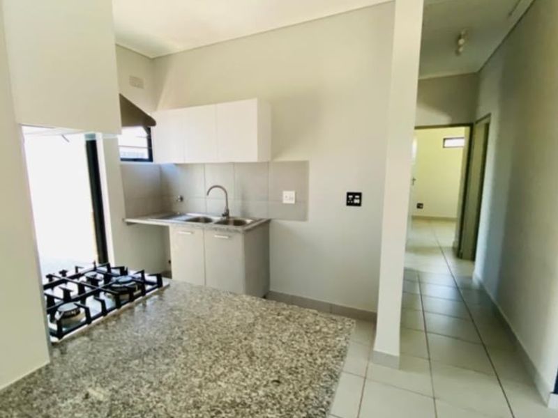 A clean, modern kitchen featuring granite countertops, a built-in gas hob with extractor, and tiled flooring, leading into a bright hallway.