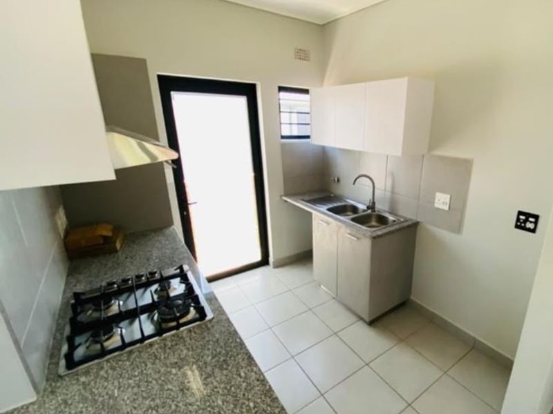 A clean, modern kitchen featuring granite countertops, a gas hob with extractor, double sink, and ample built-in cabinetry with neutral tiled flooring.