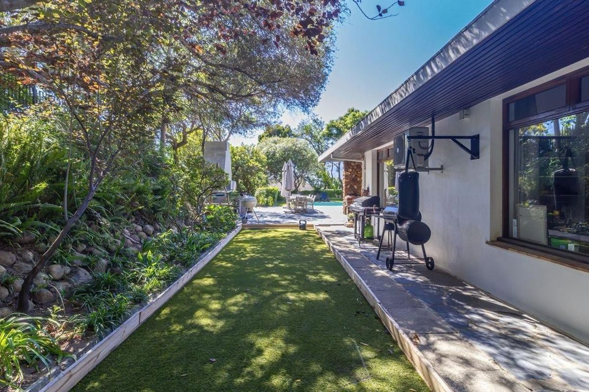 A well-maintained rear garden featuring an artificial lawn, stone retaining wall, paved walkway, and a built-in outdoor fireplace area.