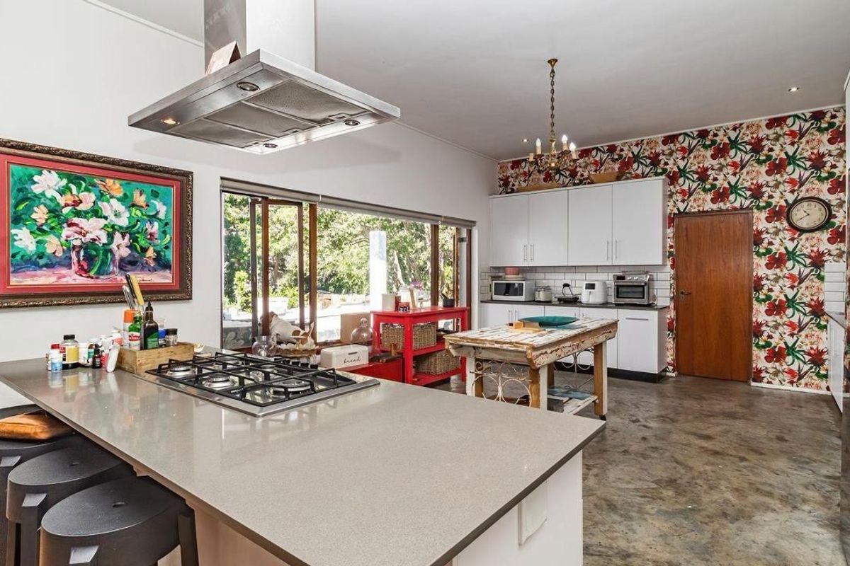 Spacious kitchen featuring a large stone-topped island with integrated gas hob, stainless steel extractor, white cabinetry, and polished concrete flooring with a feature wall.